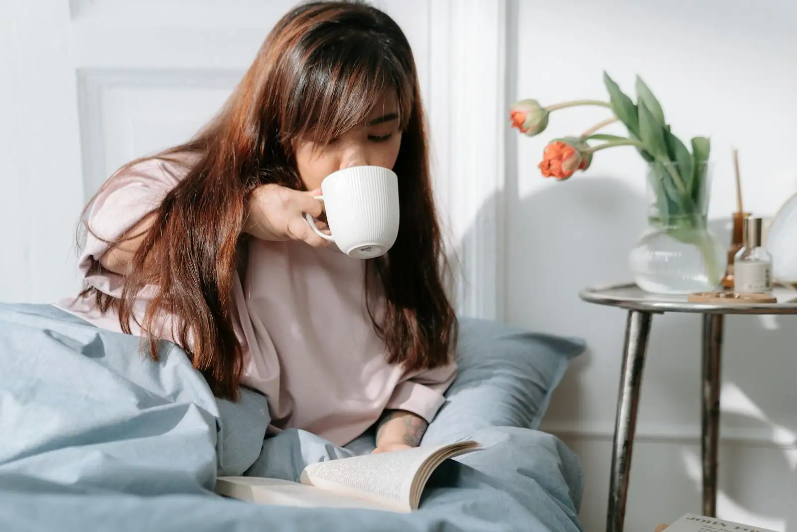 Une femme savoure un café au lit, un livre ouvert, dans la lumière douce du matin
