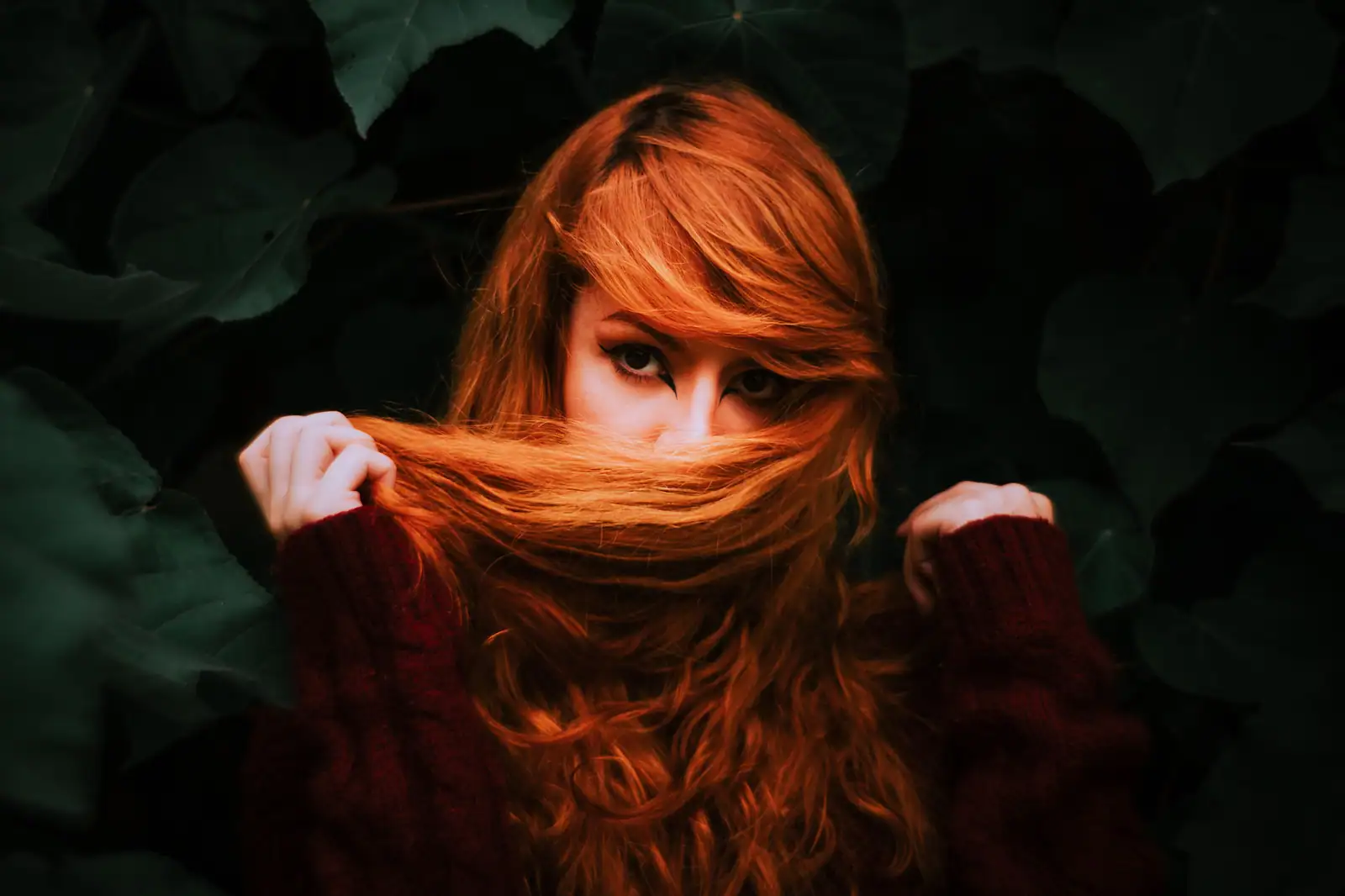 Femme aux longs cheveux d'un rouge auburn profond, en portrait sur fond de feuillage sombre