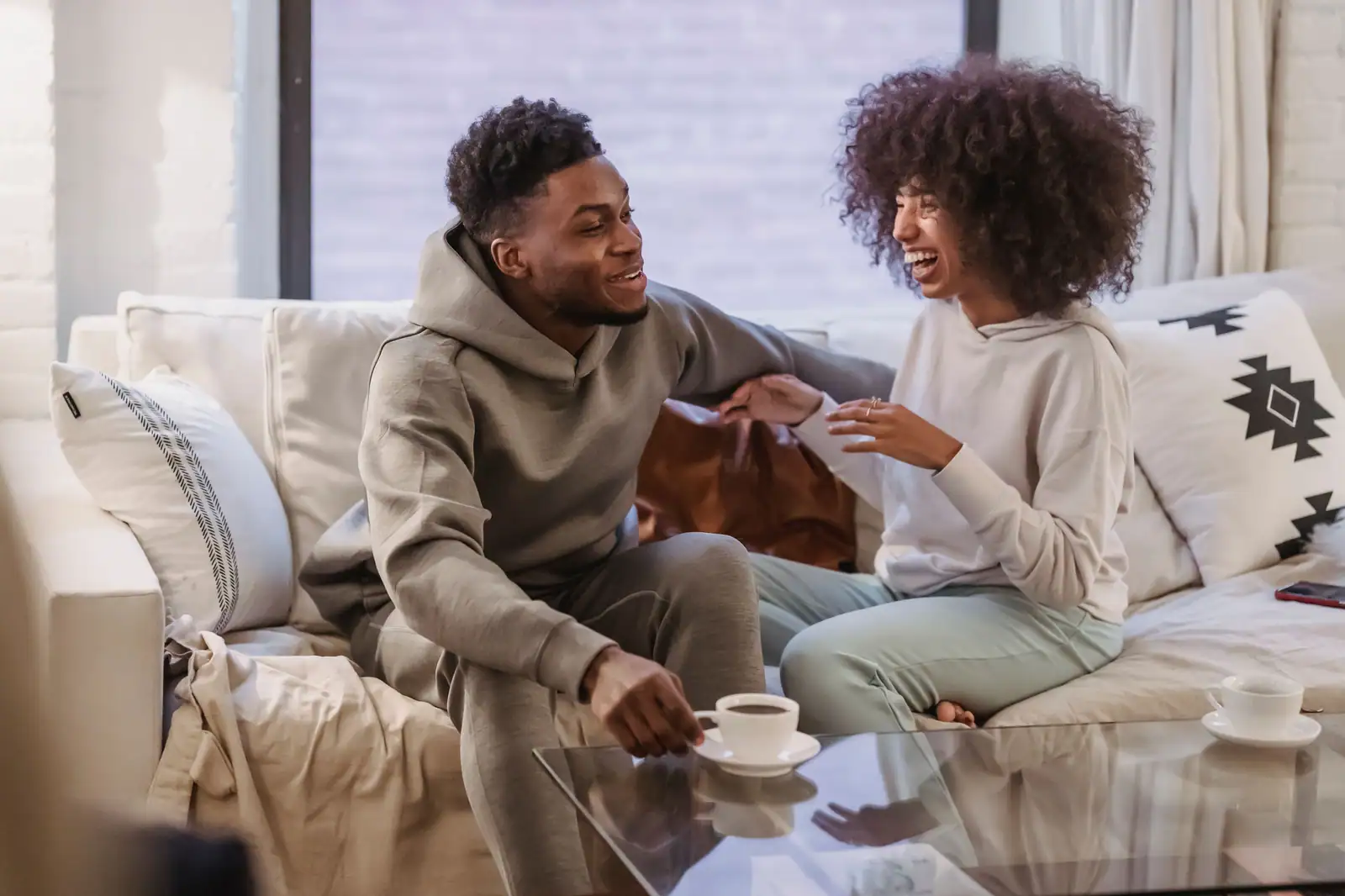 Un jeune couple souriant échange avec complicité sur un canapé, autour d'un café à la maison.