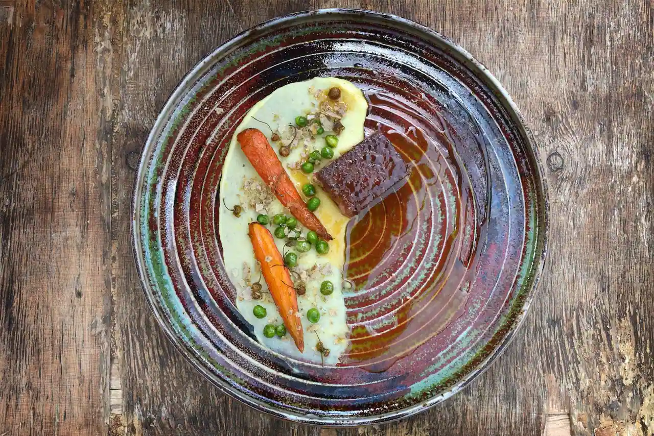 Assiette gastronomique vue de dessus : carottes glacées, petits pois et jus sur une céramique rustique