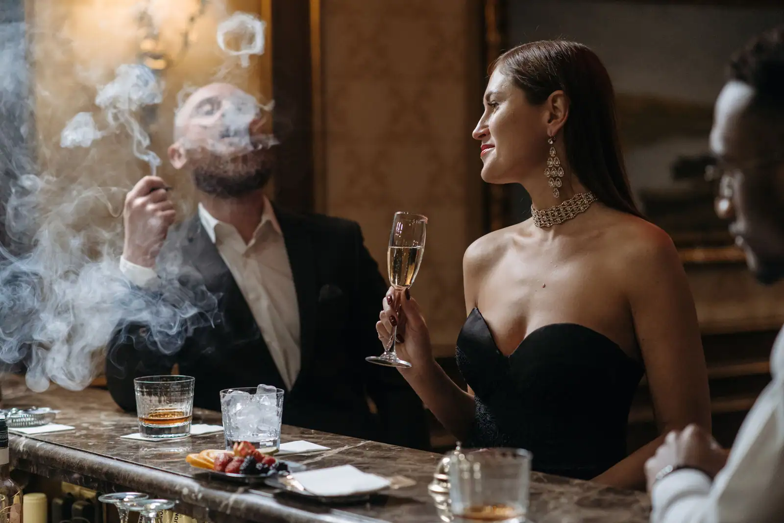 Femme en robe de soirée tenant une coupe de champagne au comptoir d'un bar feutré, un homme en smoking à ses côtés