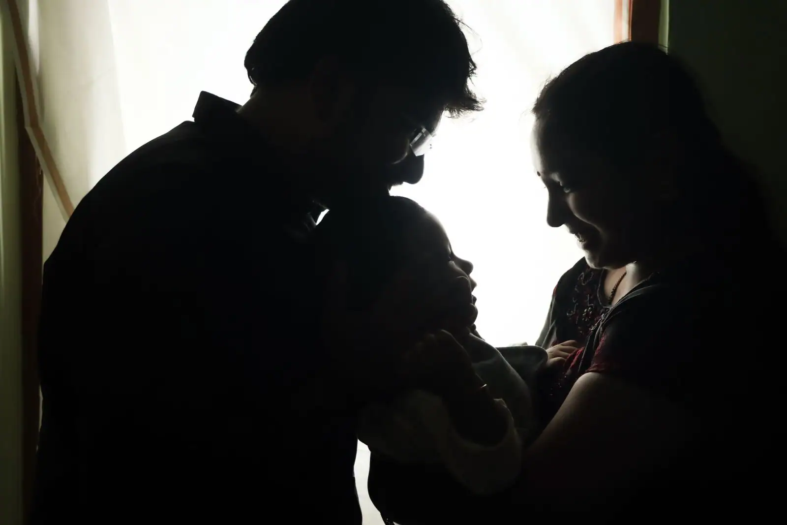 Deux parents en contre-jour tiennent tendrement leur jeune enfant devant une fenêtre lumineuse