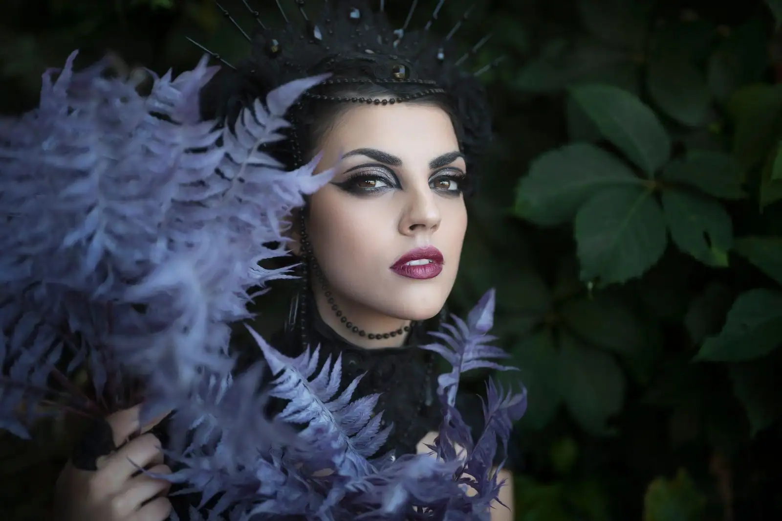 Femme au maquillage gothique : yeux charbonneux, sourcils marqués et lèvres bordeaux, coiffée d'une couronne sombre.