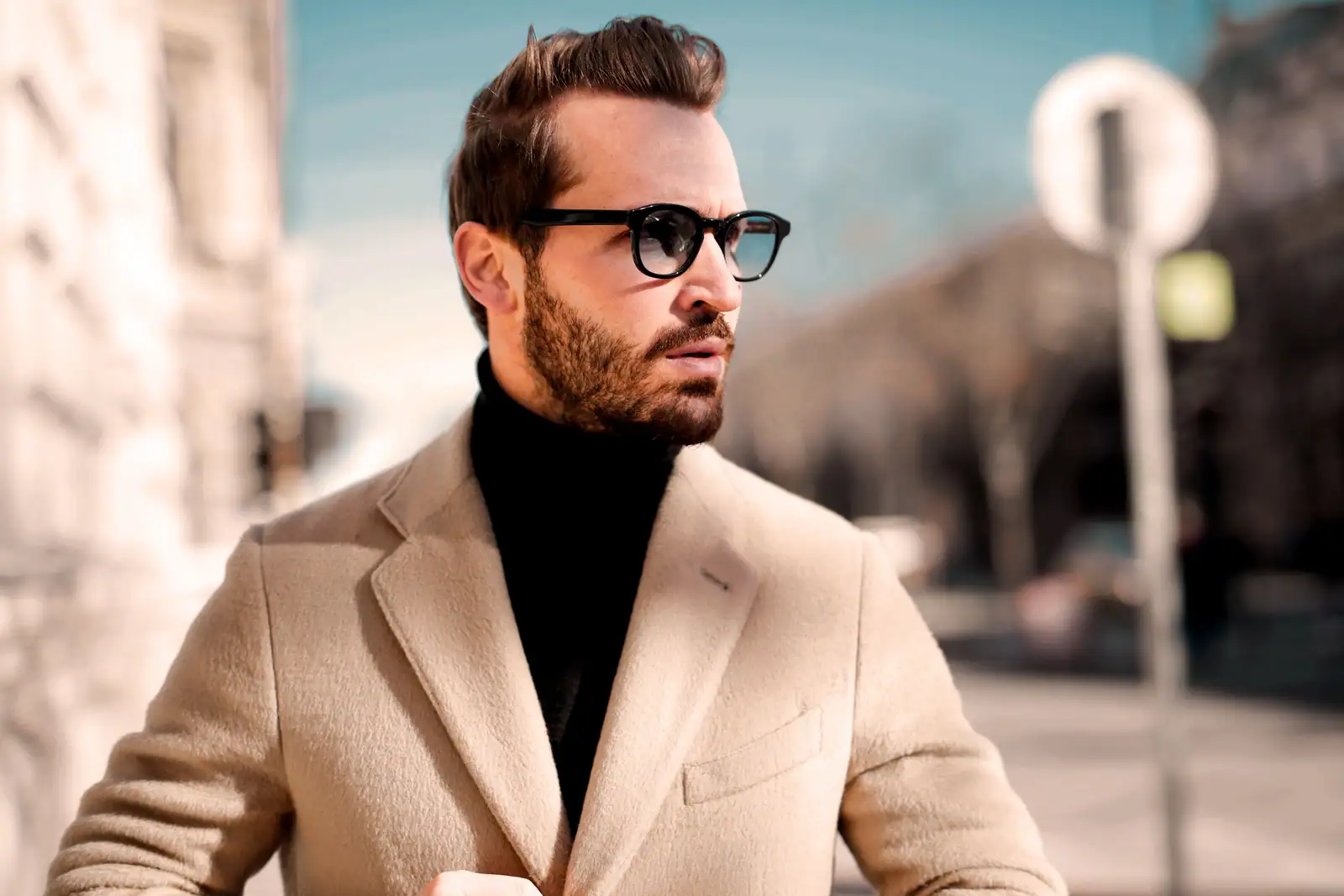 Homme élégant en manteau beige et col roulé noir dans une rue ensoleillée, mode masculine actuelle.