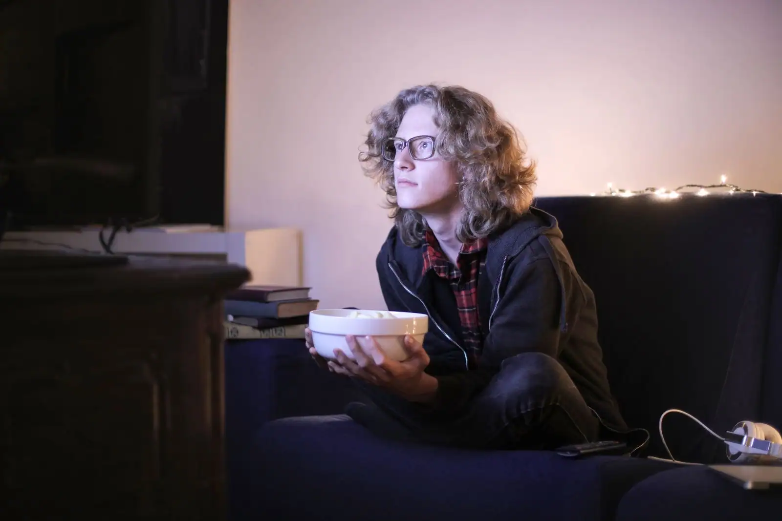 Jeune personne à lunettes dans un canapé avec un bol, regardant la télévision dans un salon en soirée