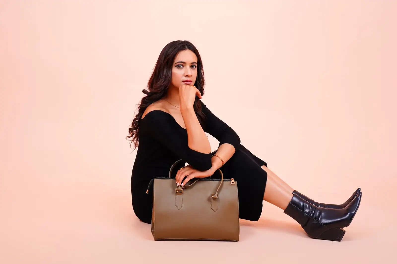 Sac à main structuré en cuir kaki posé au sol devant une femme en robe noire, sur fond rosé.
