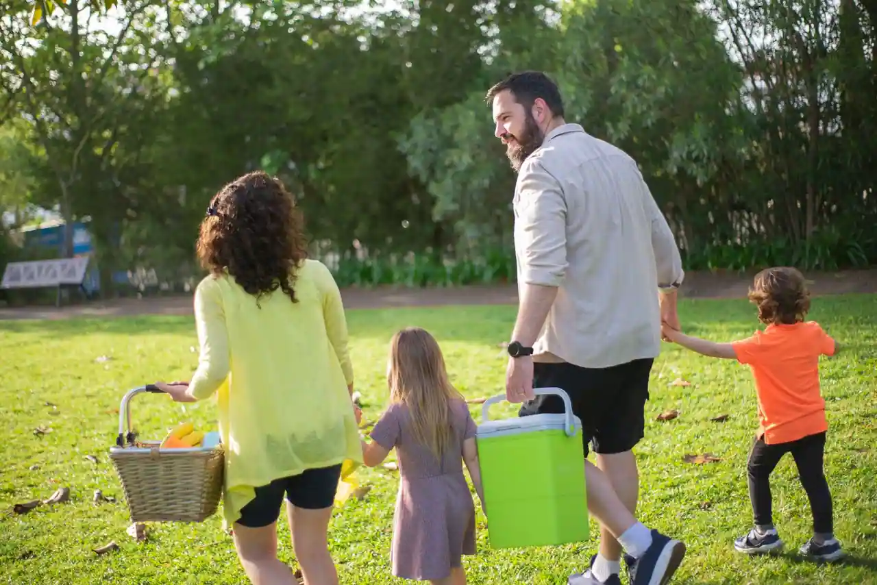 Famille avec deux enfants traversant un parc, panier et glacière en main, pour une sortie pique-nique par beau temps.
