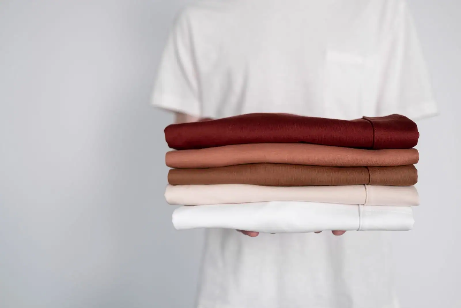 Une personne tient une pile de vêtements pliés aux teintes neutres, du bordeaux au blanc cassé.