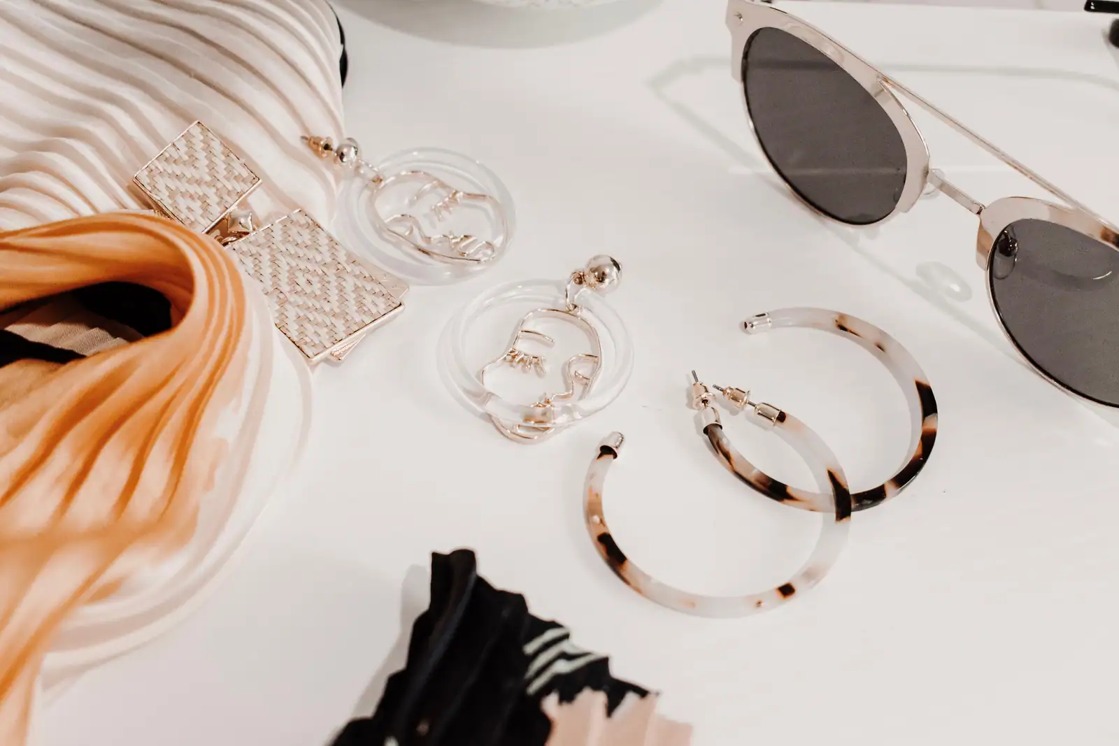 Accessoires de mode disposés à plat sur fond clair : boucles d'oreilles, foulard et lunettes de soleil