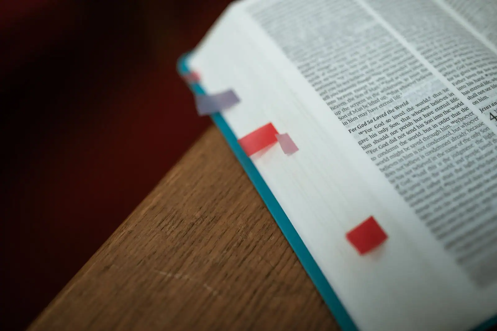 Bible ouverte en anglais posée sur le bord d'une table en bois, texte visible avec des marque-pages colorés.