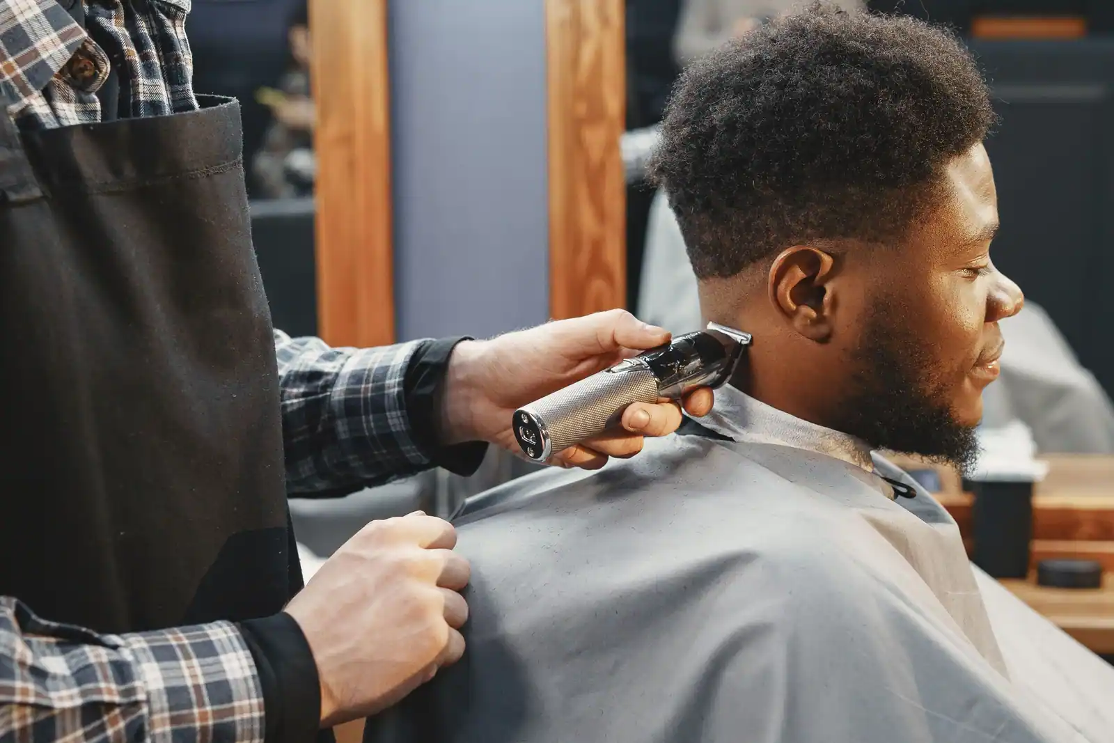 Un coiffeur dégage la nuque d'un homme barbu à la tondeuse pour réaliser un dégradé, dans un salon.