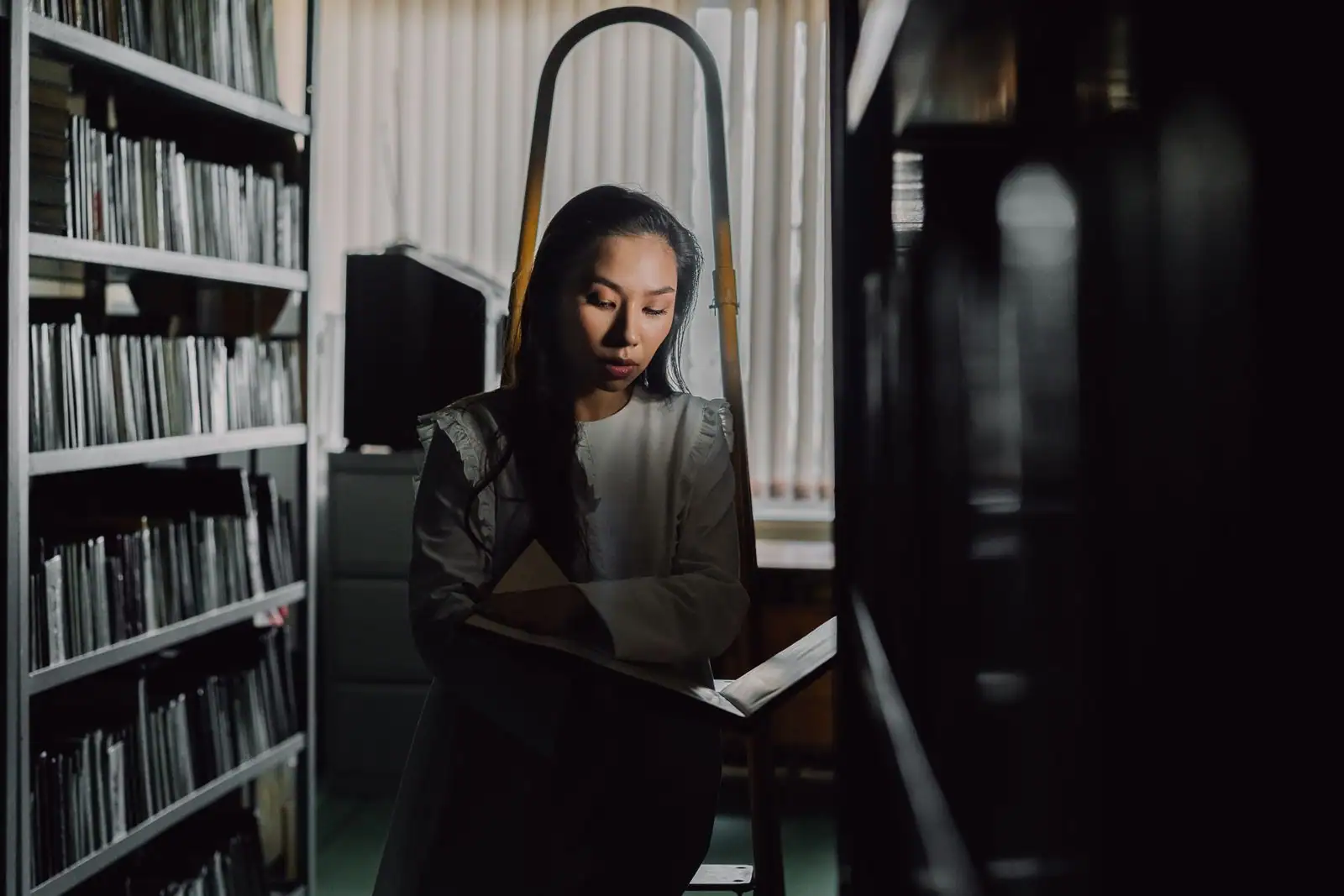 Une femme lit un livre debout entre des étagères de bibliothèque, dans une lumière douce et silencieuse.